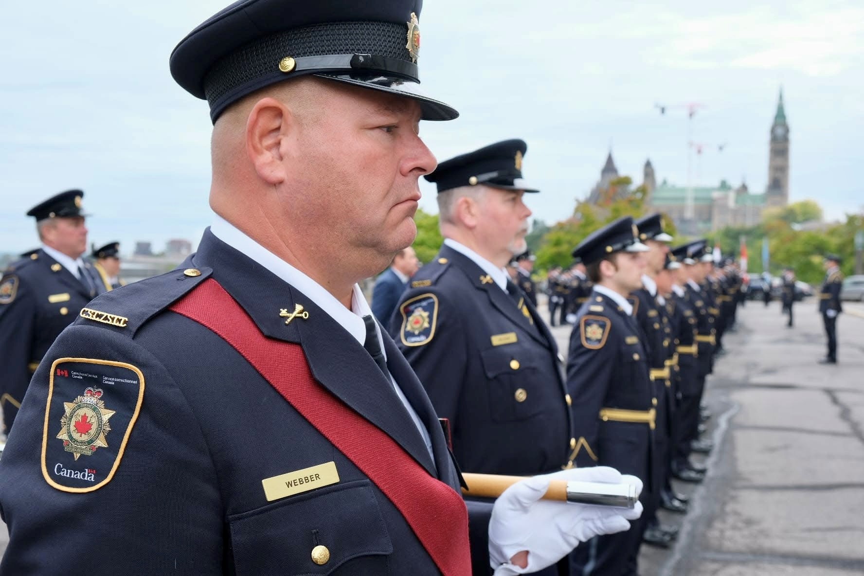 Row of uniformed CSC officers stand with Parliament Hill in backdrop.